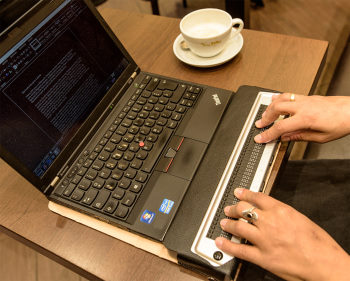 Auf dem Bild sieht man einen Laptop mit Braillezeile für bline NutzerInnen. Auf dem Bildschirm sind die ersten Zeilen eines Textes zu sehen. Neben dem Rechner steht eine Tasse Kaffee