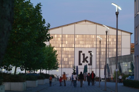 Das Bild zeigt die Fassade des Kampnagel Hamburg mit einigen Bäumen, Laternen und Gästen