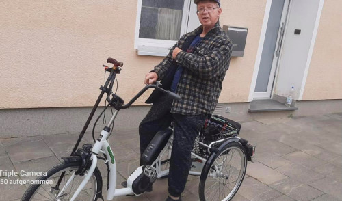 Walters Gehstock lehnt an seinem neuen weißen E-Dreirad. Er sitzt lächelnd auf dem Gefährt, welches er durch die Hochwasser Hilfe bekam