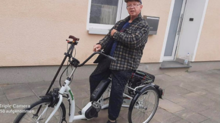 Walters Gehstock lehnt an seinem neuen weißen E-Dreirad. Er sitzt lächelnd auf dem Gefährt welches er durch unsere Hochwasser Hilfe bekam
