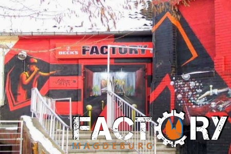 Zum Eingangsbereich der Factory Magdeburg führt eine Eisentreppe. Links und rechts sind Handläufe, das Gebäude selber ist in rot schwarz gehalten.