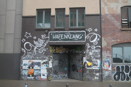 Der ebenerdige Eingangsbereich des Hafenklang Hamburg mit vielen Aufklebern, dem Logo der Location und Konzertplakaten