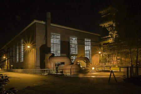 Das alte Industriegebäude der KompressorenHalle Dortmund wird nachts gelb beleuchtet. Zudem prägen aussen Kessel und Schornsteine das Bild