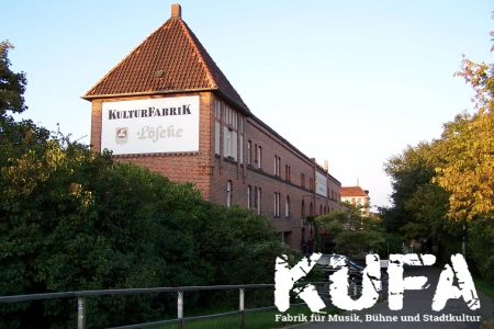 Das Gebäude der Kulturfabrik Löseke Hildesheim ist ein alter Backsteinbau, welcher von einer hohen Hecke und einigen Bäumen begrenzt wird.