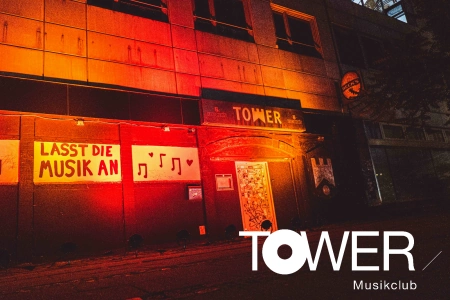 Die gelb rot beleuchtete Fassade des Tower Musikclub Bremen an der Wand neben dem Eingang sind Noten und der Spruch Lasst die Musik an auf Plakaten angebracht