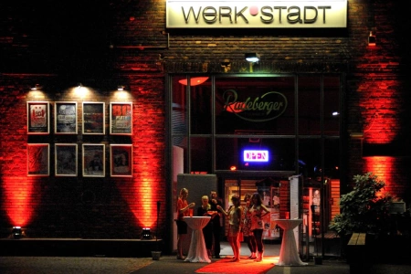Die Werkstadt Witten bei Nacht. Der Eingang ist rot beleuchtet und vor der Tür stehen einige Gäste an Stehtischen