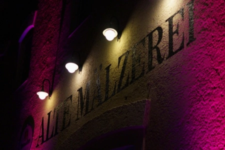 Das Schild über dem Eingang zur Alte Mälzerei Regensburg, wird von vier Lampen beleuchtet. Die Wand selber ist in ein tiefel Violett getaucht.