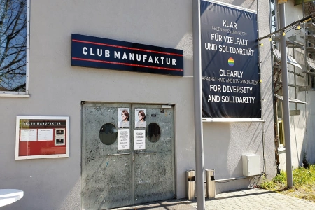 Die schwere Stahleingangstür der Club Manufaktur Schorndorf. Darüber hängt der Name der Location und rechts ein großes Transparent, welches für mehr Diversität wirbt.