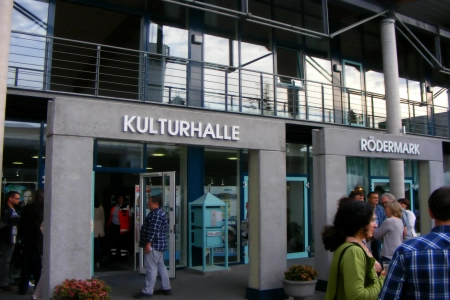 Der Eingangsbereich der Kulturhalle Rödermark besteht aus zwei großen Eingangstoren.  Über dem einen steht Kulturhalle, über dem anderen Rödermark. Einige Gäste stehen schon vor den Türen und warten