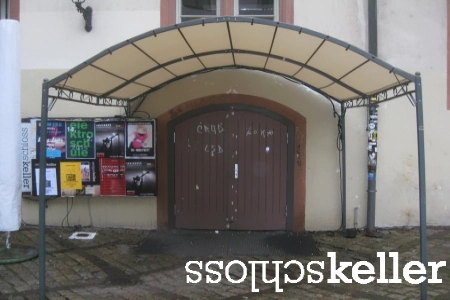 Die Holztür am Eingang des Schlosskeller Darmstadt ist mit einem Rundbogendach versehen. Daneben hängen Veranstaltungsplakate