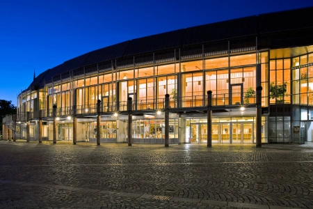 Die Stadthalle am Schloss Aschaffenburg oder Stadthalle am Schloss ist bei nacht hell beleuchtet. Die Fenster der auf alt getrimmten Fassade erstrahlen und lassen auch den Vorplatz aufleuchten