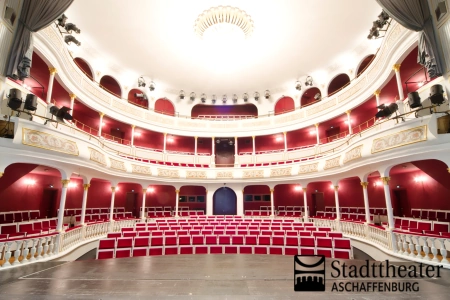 Ein Blick von der Bühne im Stadttheater Aschaffenburg direkt auf die leeren Sitze und Ränge. Edel ist alles in weiß und rot gehalten. 