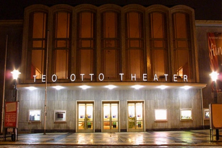 Der hell erleuchtete Eingangsbereich des Teo Otto Theater Remscheid spiegelt sich bei Nacht auf dem vom Regen nassen Vorplatz