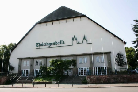 Die Thüringenhalle Erfurt von aussen, davor stehen einige Flaggenmäste und einige Büsche.