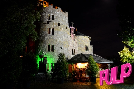 Das Eventschloss Pulp Dusiburg ist in der Nacht hell erleuchtet. Der Eingang ist überdacht und einige kleinere Bäume stehen vor dem Schloss