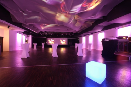Die Eventhalle Westpark Ingolstadt von innen in lila Licht getaucht. Vereinzelt stehen Leuchtwürfel und Tische im Saal verteilt.