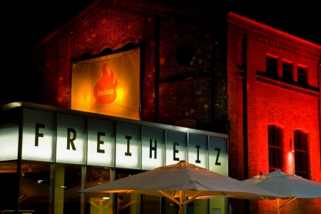 Das Freiheiz München ist bei Nacht Rot / Orange /gelb beleuchtet. Über dem Eingang hängt eine große Leuchtreklame der Location und davor steht ein Sonnenschrim