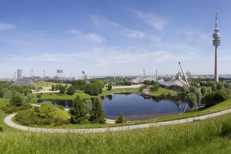 Der Olympiapark umschließt das Gelände der Olympiahalle München. Mittig ist der große See zusehen, an ihm führt ein Rundweg um das Gelände. Rechts erkennt man den Olympiaturm und links liegt die Olympiahalle, sowie das Stadion