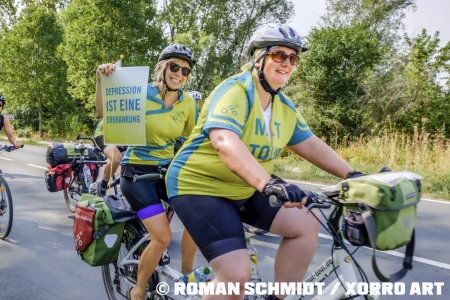 Die Mut-Tour 2022 Zwei Frauen sind in Fahrradkleidung auf einem Tandem unterwegs. Die hintere hält ein Schild mit der Aufschrift Depression ist eine Erkrankung
