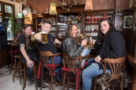 Die vier Bandmitglieder von Tankard sitzen an der Bar und halten ein Glas Bier in die Kamera. Alle vier lachen oder lächeln