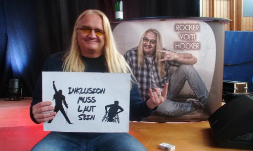 Der Rocker vom Hocker hält ein Schild mit der Aufschrift Inklusion Muss Laut Sein und zeigt die Pommesgabel