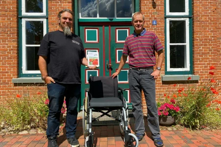 Ron und der Pastor der Gemeinde Süderhastedt stehen vor dem Pfarrhaus mit einem neuen Rollstuhl
