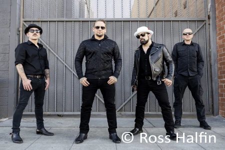 Die vier Musiker von Volbeat stehen nebeneinander an einem Tor. Alle vier sehen direkt in die Kamera und haben lässig die Hände in den Taschen