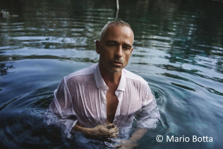 Der italienische Sänger Eros Ramazzotti steht mit einem halb geöffneten weißen Hemd bis zur Brust in einem See. Sein Blick schweift ins Leere. Die Hände hat er vor Brust und Bauch.
