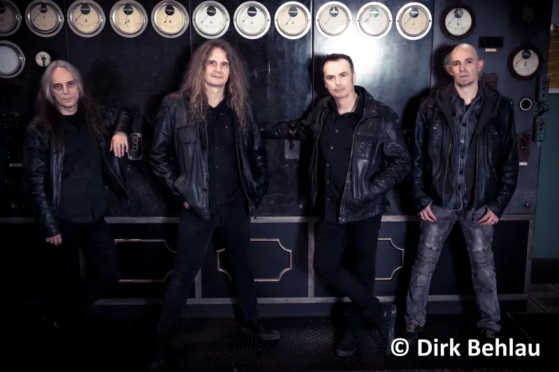 Die Musiker von Blind Guardian stehen vor einer alten Maschine mit runden Ableseskalen. Alle vier Musiker sind lässig angelehnt und schwarz gekleidet. Der Gesichtsausdruck ist neutral.