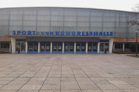 Der Vorplatz der Sport- und Kongresshalle Schwerin ist mkt großen Betonplatten ausgelegt. Über dem Eingang der Halle prankt der Name der Location in blauen Buchstaben.
