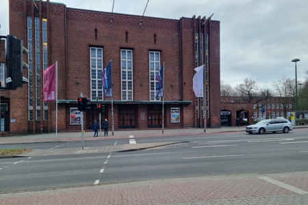 Eine breite Hauptstraße befindet sich vor den Eingängen zur Veranstaltungsstätte Deutsches Haus Flensburg. Vor dem Gebäude wehen vier Flaggen