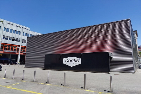 Das Les Docks Lausanne hat eine modere Fassade und einen großen Schriftzug mit dem Namen Docks. Davor stehen einige Avsperrpoller.