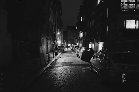 Eine nur spärlich beleuchtete Straße in Schwarz-Weiß gehalten. Es ist tiefste Nacht, nur eine einzelne Person ist schemenhaft in der Ferne zu sehen.
