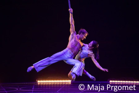 Ein männlicher und ein weiblicher Akrobat des Cirque du Soleil, hängen engumschlungen an einem Band. Der Mann hält sich dabei an nur einer Hand. Die Frau ist an ihn geklammert und lässt ihren Arm hängen.